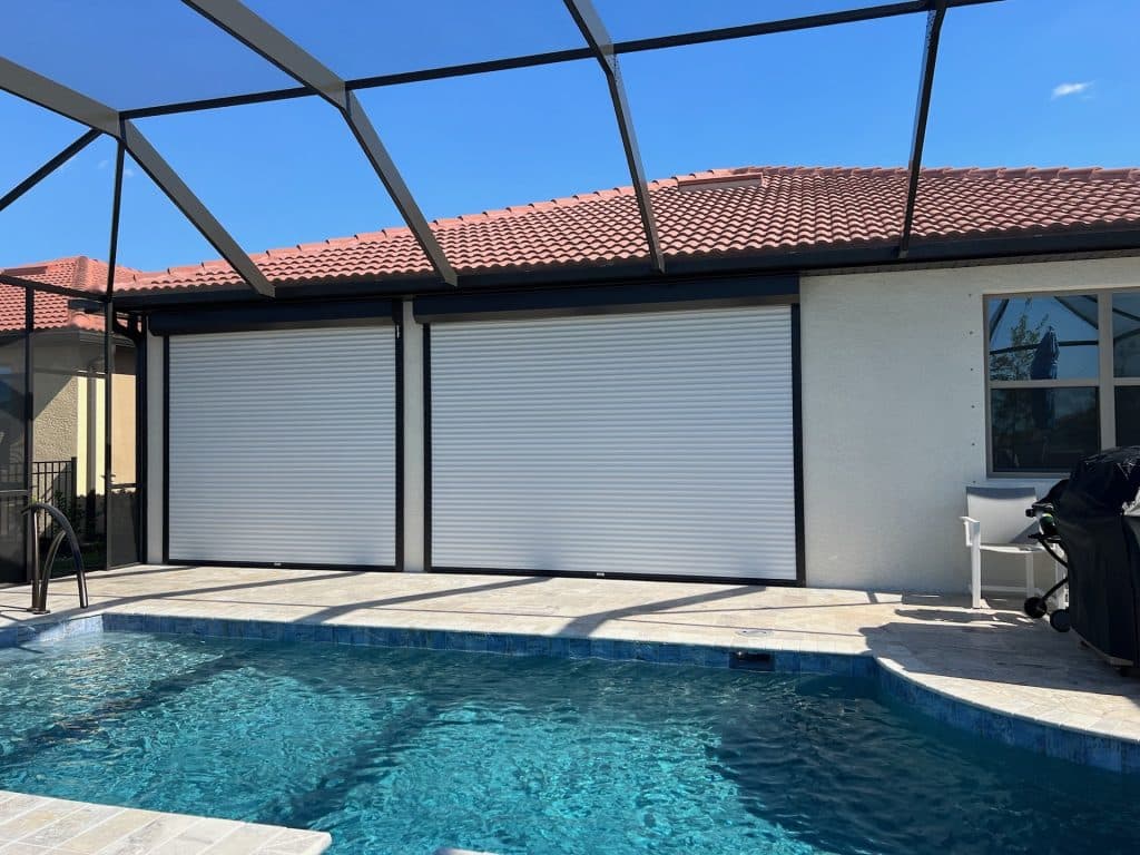 A backyard pool with screen enclosure, two white roll-down shutters