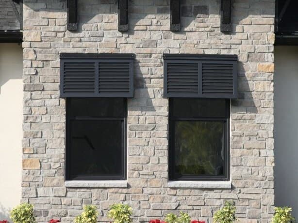 Two windows with black aluminum Bahama shutters on a stone exterior wall.